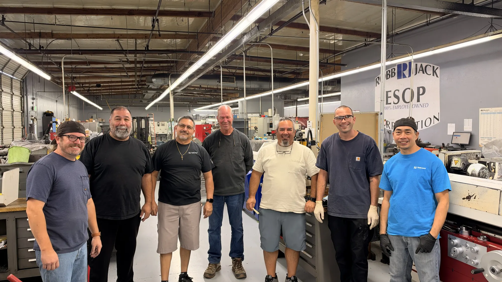 Seven smiling RobbJack team members standing together on the grinding floor in Lincoln, California, under a wall sign that reads RobbJack ESOP, 100% employee owned.