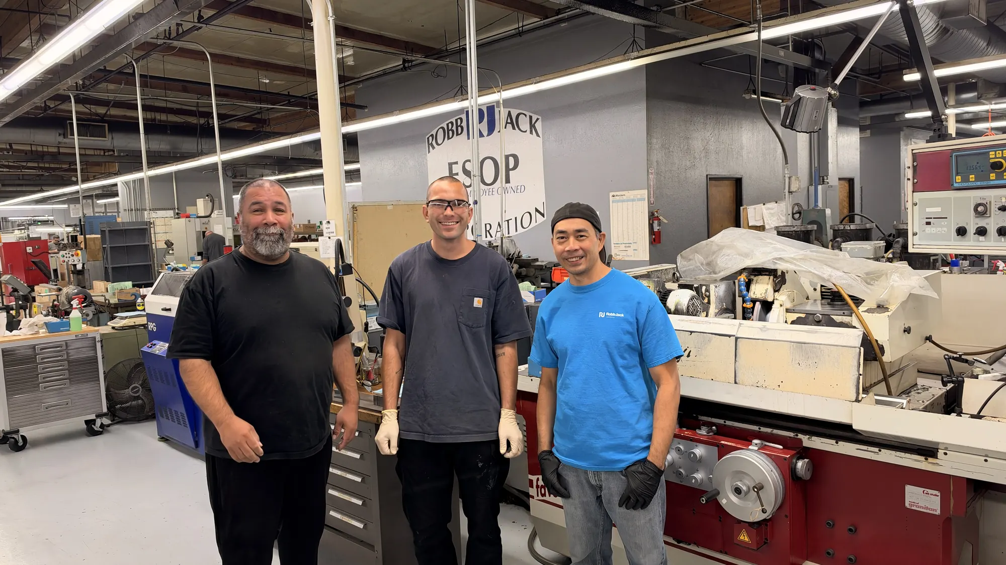 Three RobbJack team members smiling beside a CNC tool grinder in the Lincoln shop.